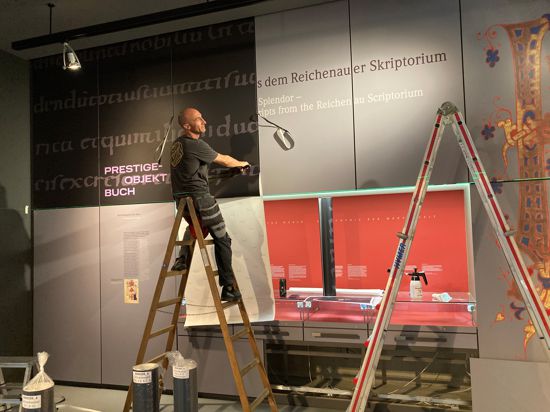 Ein Mann auf einer Leiter bezieht eine hohe Wand mit einer schwarzen Folie, die mit mittelalterlichen Schriftzeichen bedruckt ist