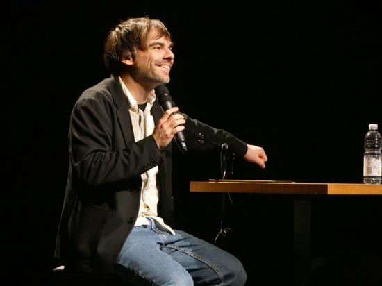Mann auf Bühne mit Mikro in de rechten Hand, mit der linken klopft er auf einen Bistrotisch 