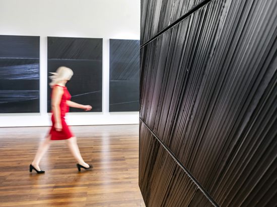 Eine blonde Frau in totem Kleid geht schnellen Schritts durch eine Ausstellung mit schwarzen Bildern.