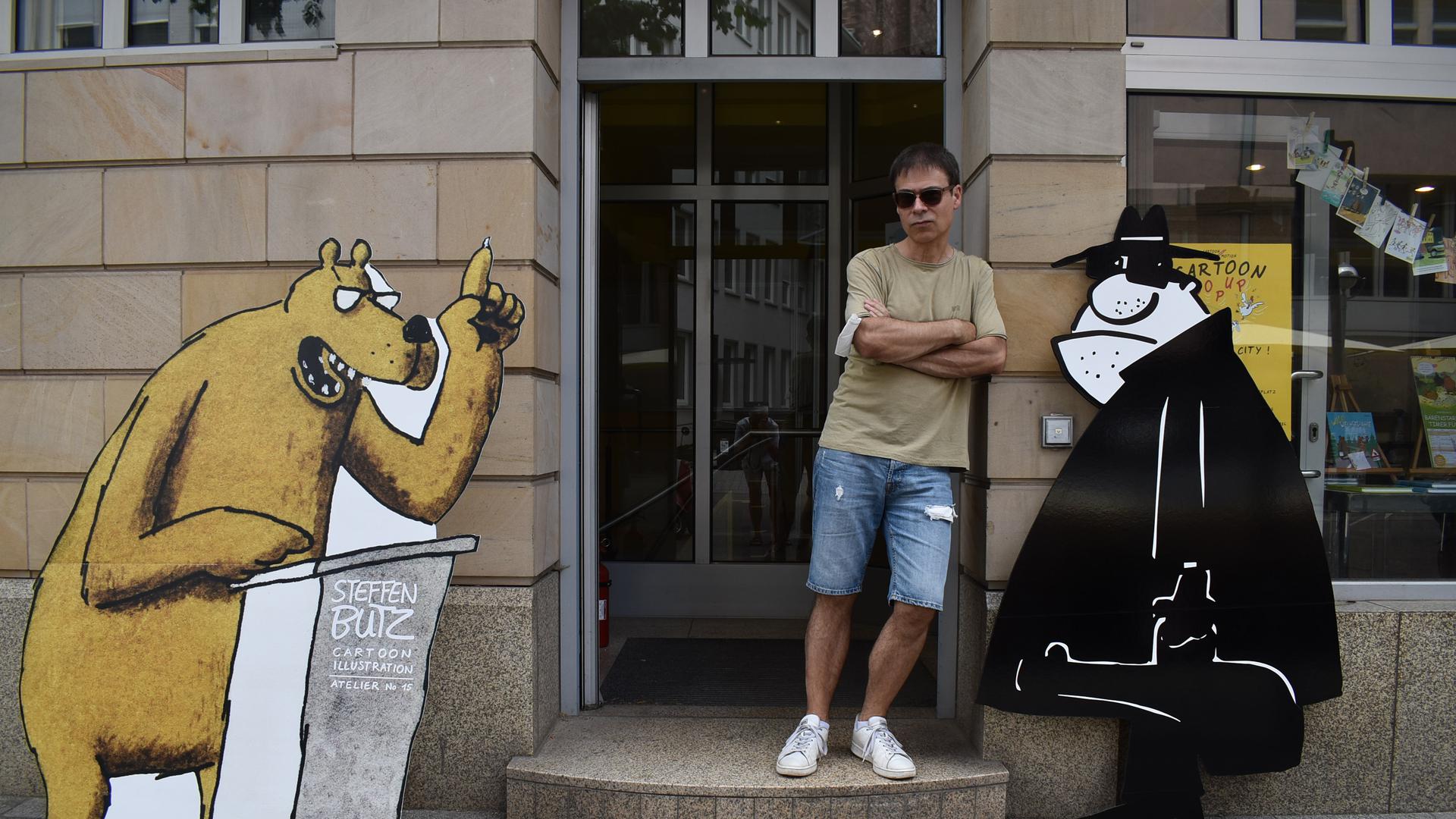 Steffen Butz neben zwei großen Cartoonfiguren in einem Hauseingang. 