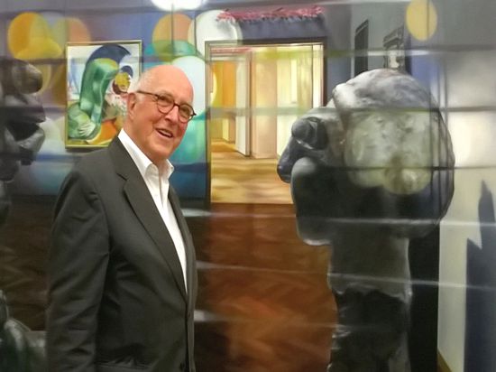 Ein Mann mit Brille, Anzug, weißes Hemd, steht vor einer Malerei, in der sich mehrere Kunstwerke zu spiegeln scheinen.