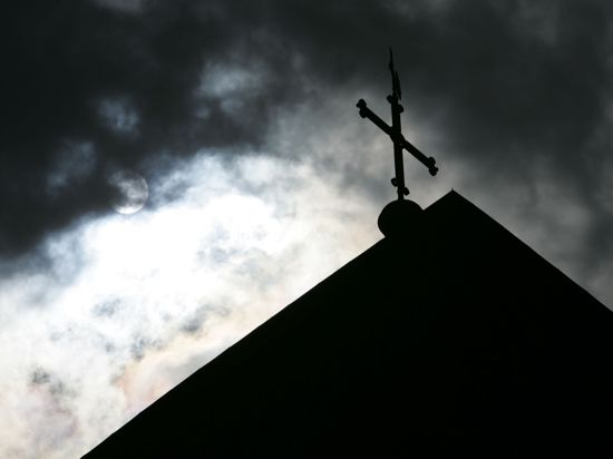 Im Gegenlicht und vor wolkenverhangenem Himmel ist die Kirchturmspitze eines Doms mit Kreuz zu sehen.