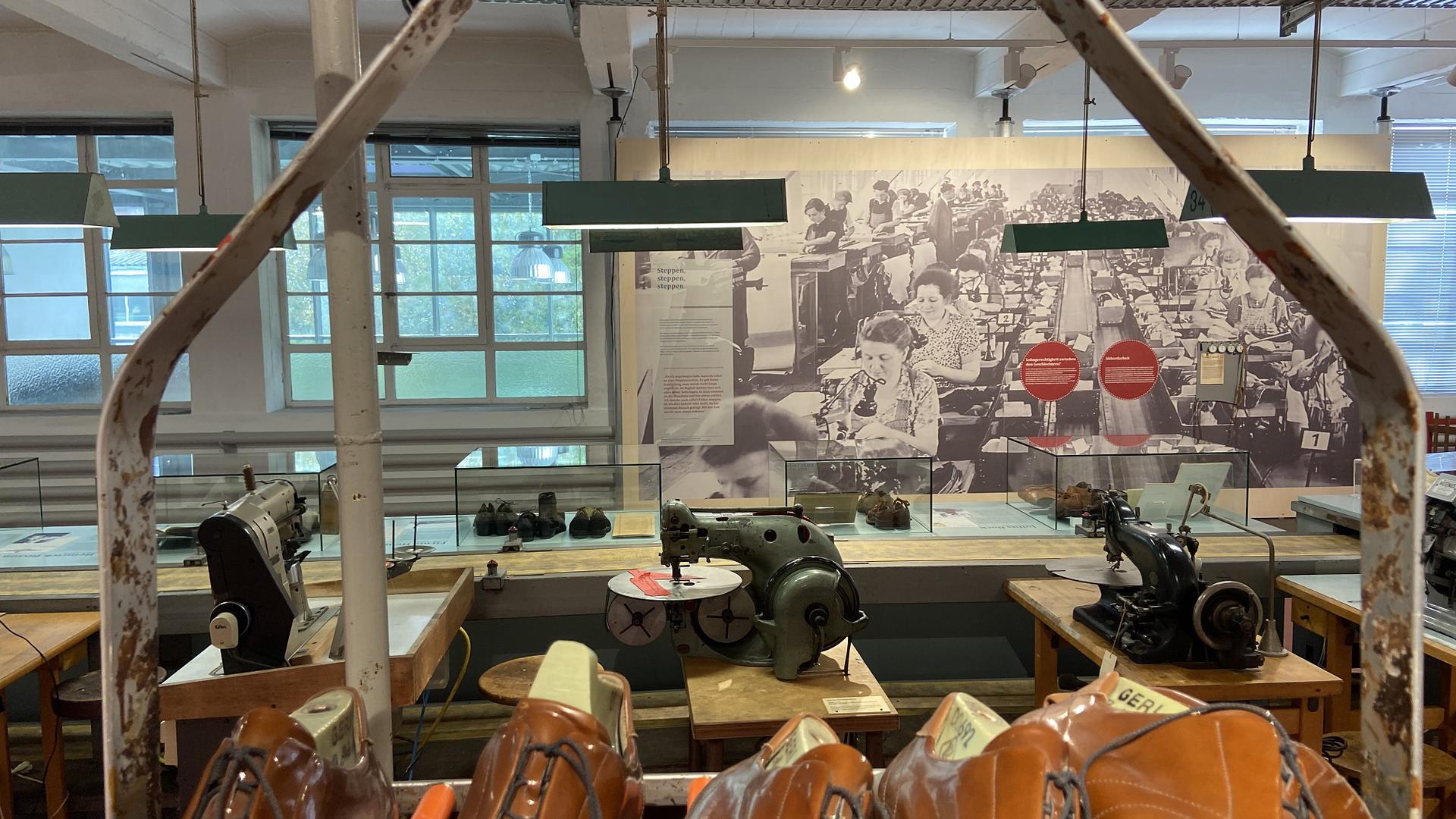 Nachgebaute Näherei einer Schuhfabrik. Im Hintergrund hängt ein große historische Aufnahme an der Wand, die eine Fabrikhalle mit zahlreichen Arbeiterinnen an Nähmaschinen zeigt.