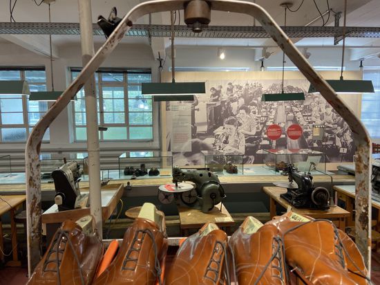 Nachgebaute Näherei einer Schuhfabrik. Im Hintergrund hängt ein große historische Aufnahme an der Wand, die eine Fabrikhalle mit zahlreichen Arbeiterinnen an Nähmaschinen zeigt.