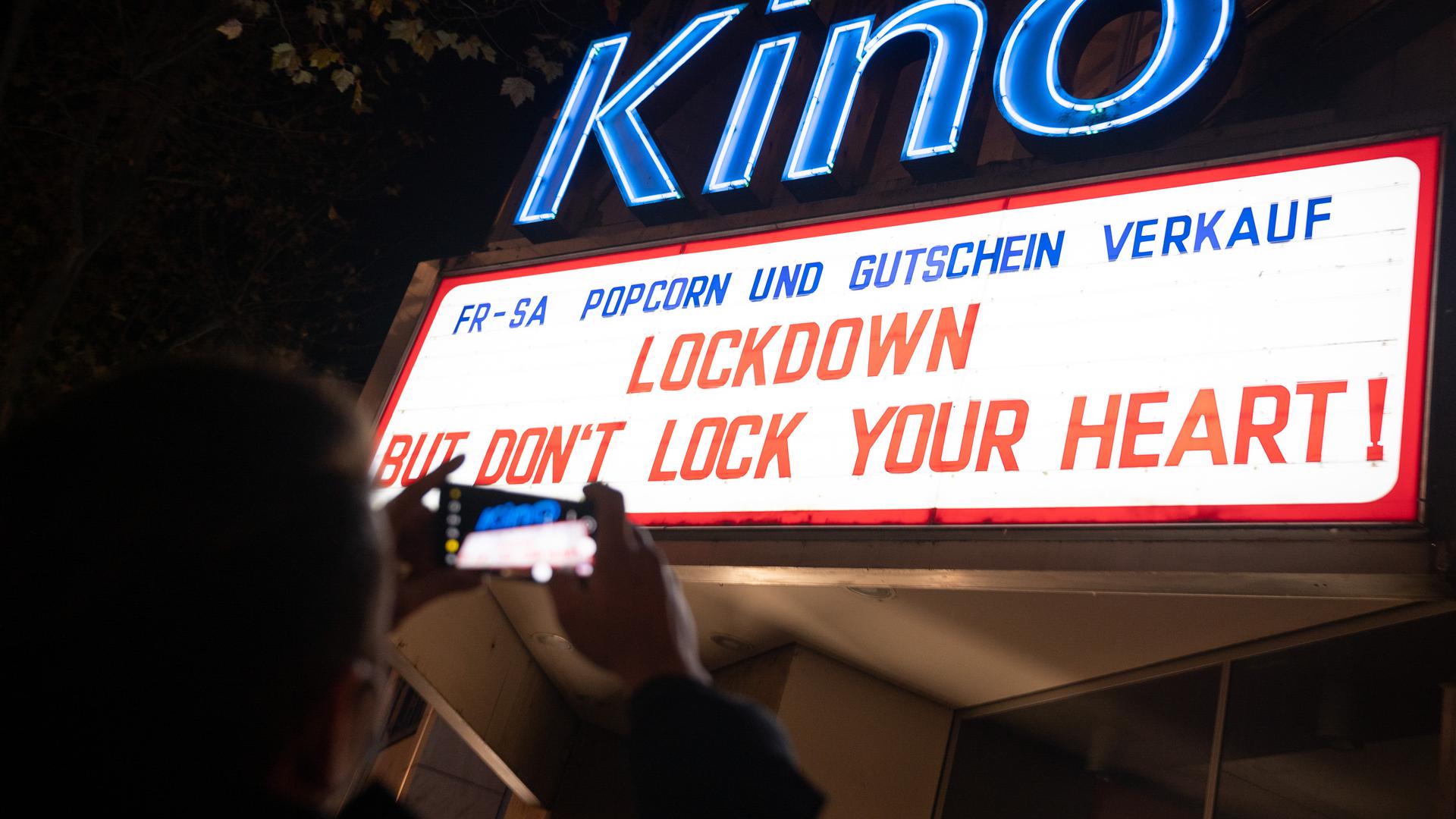 Ein Mann fotografiert die beleuchtete Anzeigetafel eines Kinos auf der "Lockdown but don't lock your heart!" steht. 