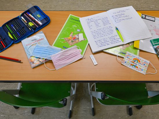 Unterrichtsmaterialien und Masken liegen während einer Pause auf einem Tisch in einem Klassenraum einer Grundschule. 