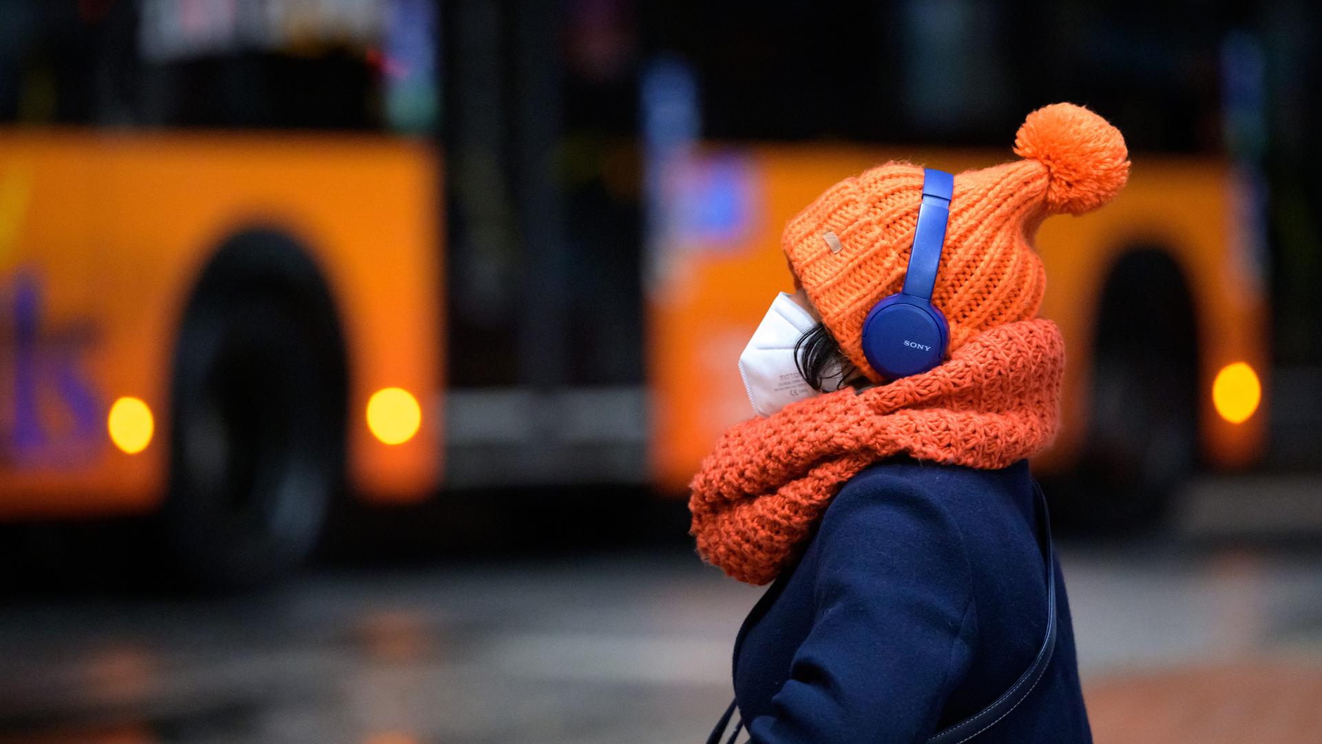 Eine Frau sitzt mit FFP2-Maske, dicker Mütze und Kopfhörern an einem Busbahnhof, während ein Linienbus vorbeifährt. 