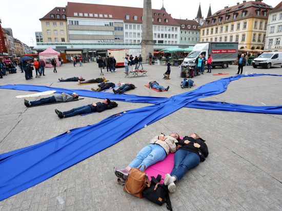Menschen liegen im Rahmen einer Liegend-Demonstration zum internationalen ME/CFS-Tag entlang einer als Schleife gelegten Stoffbahn. 