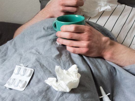 Ein Mann liegt mit Taschentüchern, Teetasse, Nasenspray und Tabletten im Bett. Ähnlich wie bei Corona kann auch eine Grippe-Infektion bei Klinikpatienten schwerwiegende Langzeitfolgen haben. 