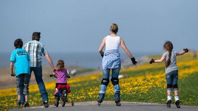  Ein Mann, eine Frau und drei Kinder fahren mit Inline-Skates, Fahrrad und Tretroller durch Wiesen.