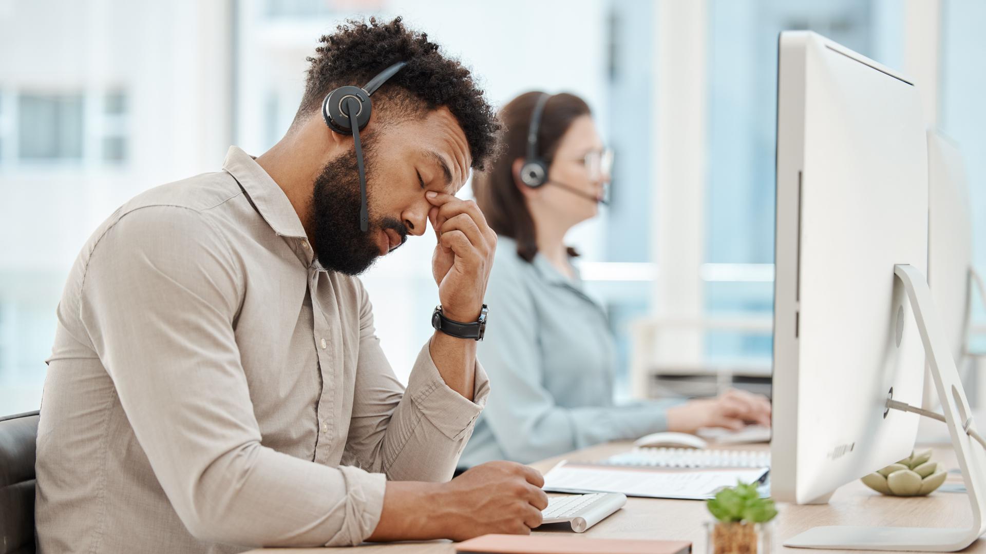 Ein Mitarbeiter im Call-Center sitzt offensichtlich müde und gestresst vor dem Computer. Er hat die Augen geschlossen, den Kopf hält er gesenkt und massiert sich mit den Fingern die Nase-Stirn-Partie.