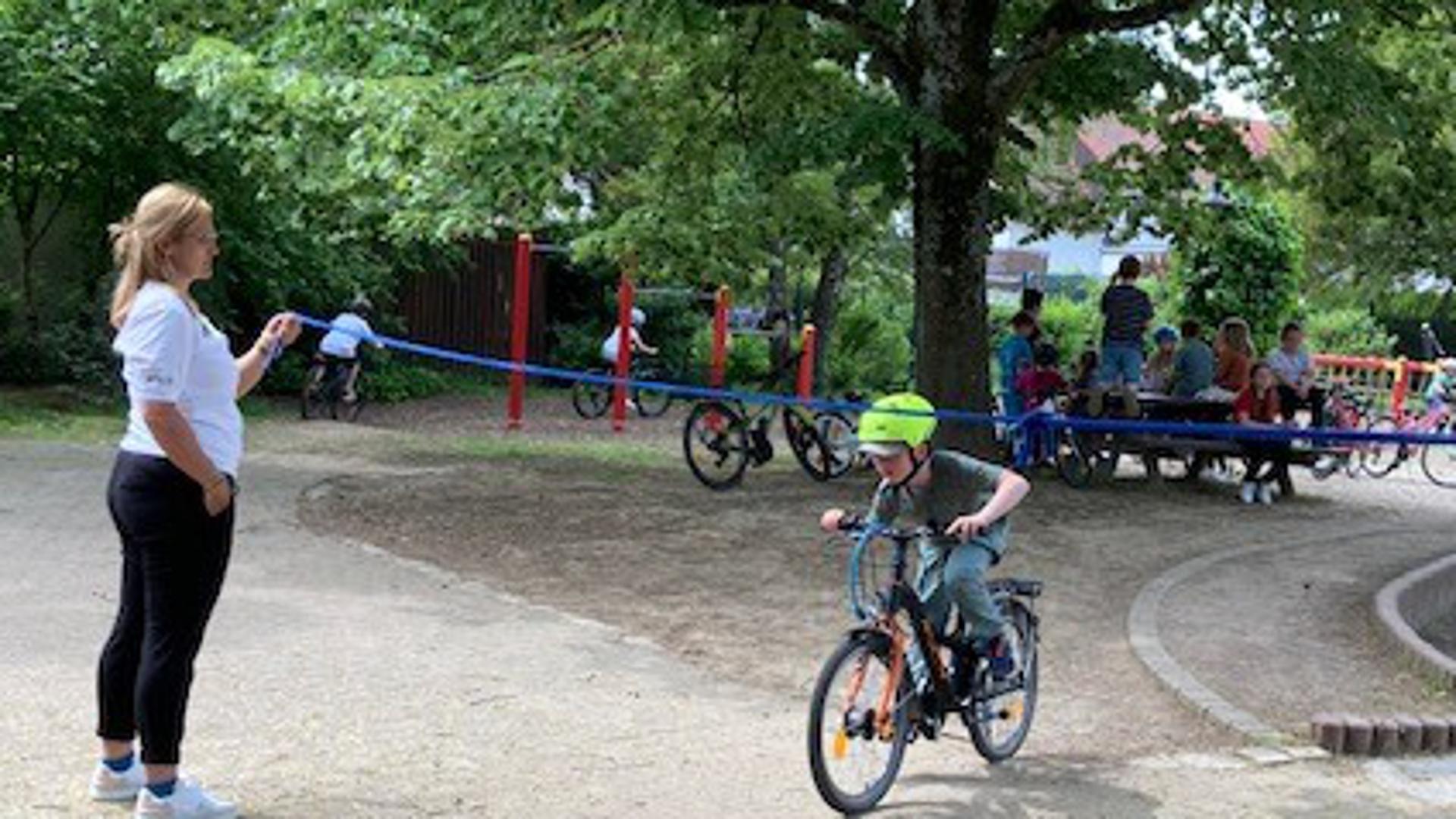 Ein Kind auf einem Fahrrad fährt unter einem Seil durch, das von zwei Betreuerinnen gehalten wird. 