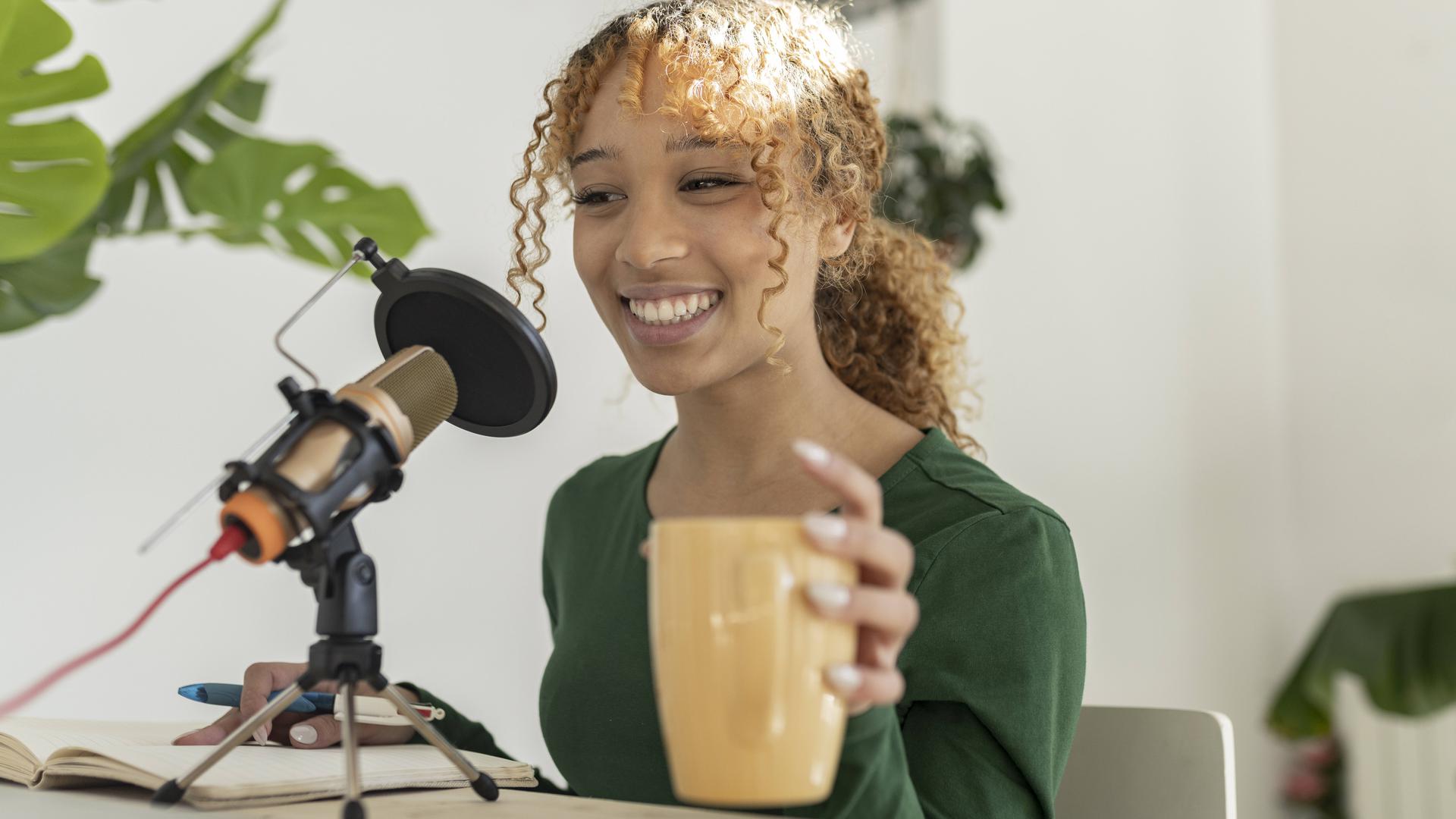 Smiling young ethnic female recording podcast on laptop with professional microphone while sitting at table with glass of fresh smoothie and notebook, Model released, Property released JoseCarlosIchiro_WomenAtHome_56.jpg Copyright: xJosexCarlosxIchirox