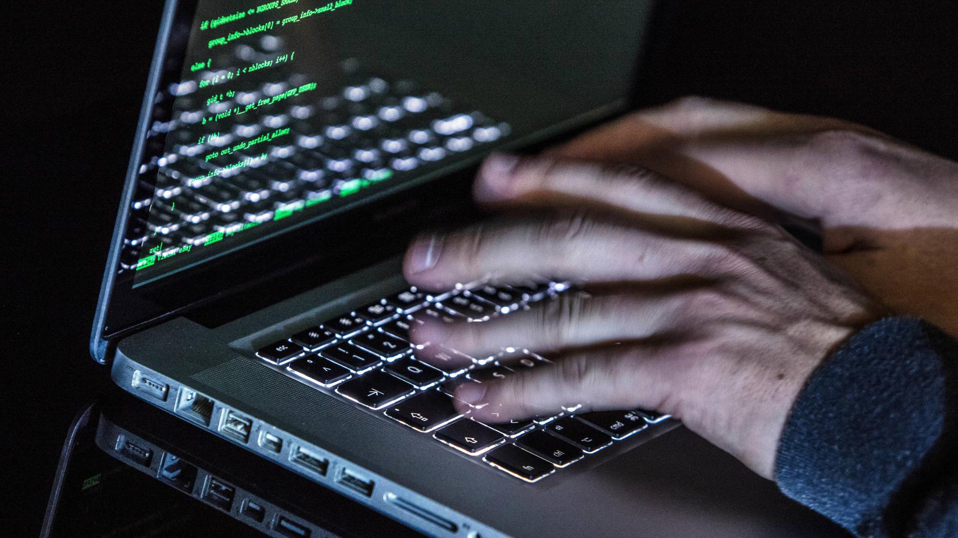 Ein Mann tippt auf einer beleuchteten Tastatur eines Laptops. Auf dem Monitor ist ein Hacker-Programm geöffnet. 