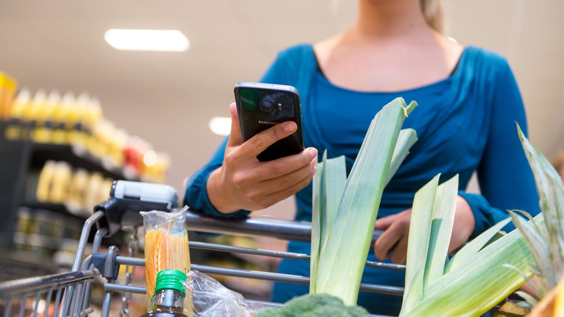 Eine Frau im Supermarkt schiebt einen Einkaufswagen und hat ein Handy in der Hand. 