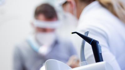 Ein Zahnarzt und eine Zahnarzthelferin mit Mund-Nasen-Schutz und Schutzvisieren versorgen eine Patientin in einer Praxis. 