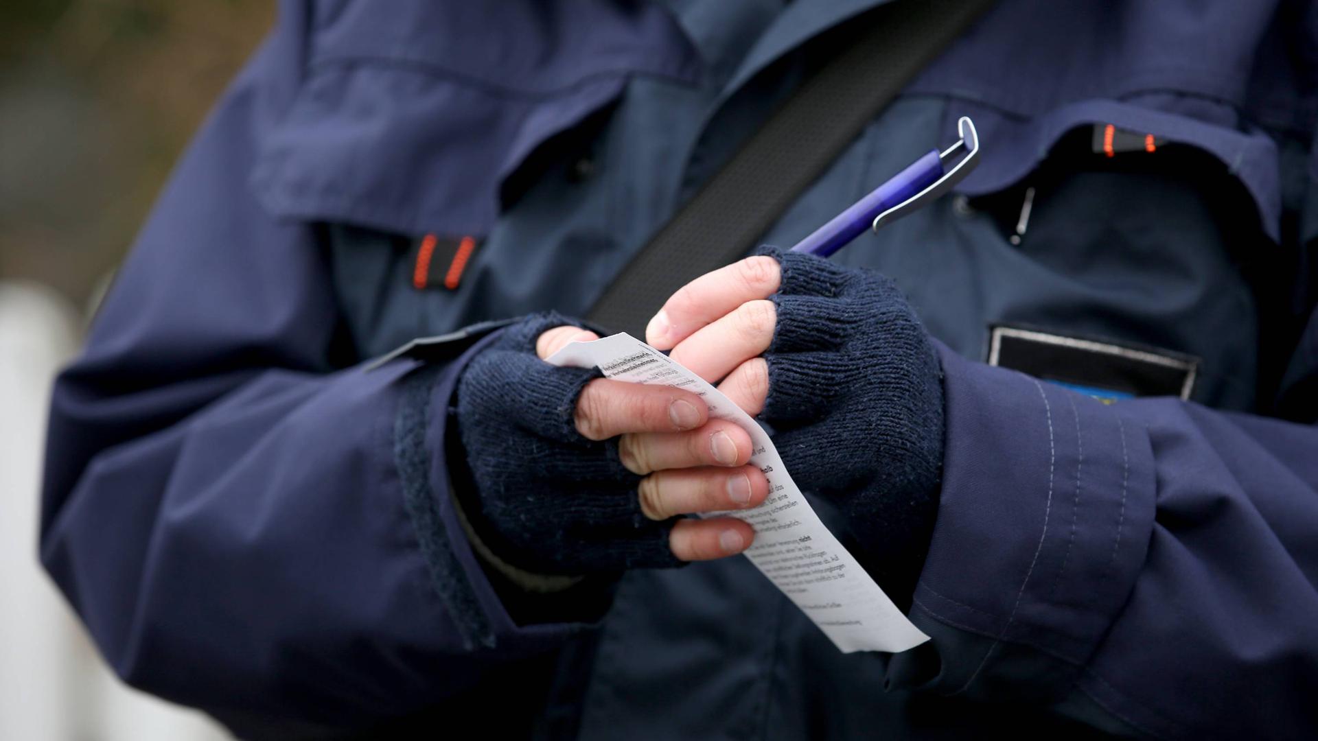 Eine Person in blauer Polizeijacke schreibt einen Strafzettel. Der Bildausschnitt ist auf die Hände begrenzt, die in fingerlosen, dunklen Handschuhen stecken.