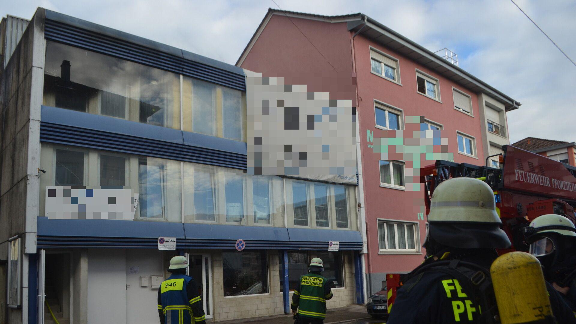 Front eines Mehrfamilienhauses in Pforzheim. Aus der oberen linken Wohnung mit Balkon dringt Rauch. Vor dem Haus sind drei Feuerwehrleute zu sehen.