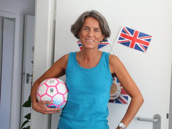 Eine Frau steht mit dem Ball unterm Arm vor einer Tür, an der eine englische Flagge hängt.
