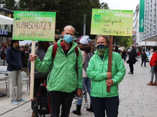 Ein Mann und eine Frau in grünen Jacken und mit Atemschutz halten zwei Schilder hoch: „Klimaschutz jetzt und für alle“ sowie „Nachhaltigkeit statt Profitgier“ steht darauf, sowie jeweils „Greenpeace Pforzheim“.