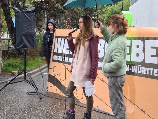 Grüne Jugend Sprecherin, Sarah Heim mit Mikrophon. Im Hintergrund die weiße Mauer der Abschiebehaftanstalt.