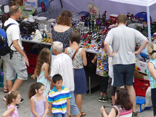 Mehrere Menschen stehen vor einem Flohmarktstand. Auf dem langen Tisch sind verschiedene Artikel wie Lego und Geschirr aufgebaut.