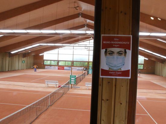 Die nahezu leere Tennishalle der PSG Pforzheim, drei Felder mit rotem Granulat. Im Hintergrund trainiert jemand, vorne ist ein Schild angebracht mit einem Hinweis, Maske zu tragen.