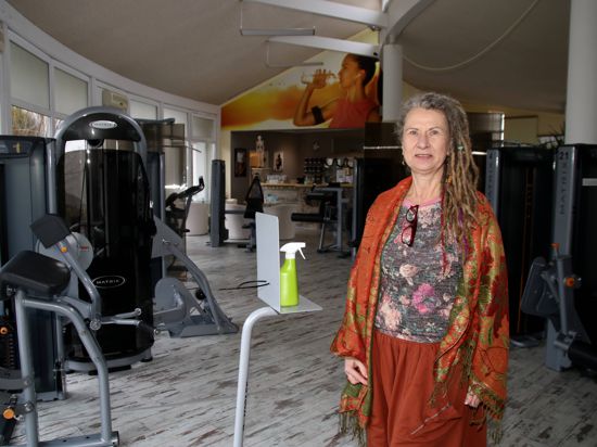 Frau mit Rasta-Locken und buntem Gewand steht inmitten von Fitnessgeräten in einer leeren Trainingshalle.