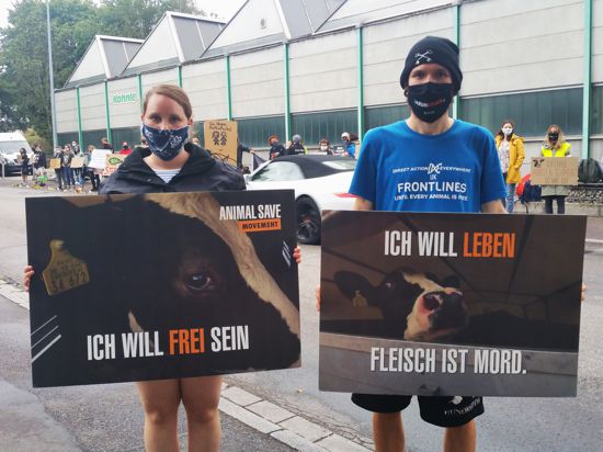 Zwei Demonstranten stehen mit Mundschutz und Schildern im Vordergrund. Auf den Schildern steht „Ich will frei sein” und „Ich will leben. Fleisch ist Mord”. Im Hintergrund sind weitere Demonstranten vor einer Halle.