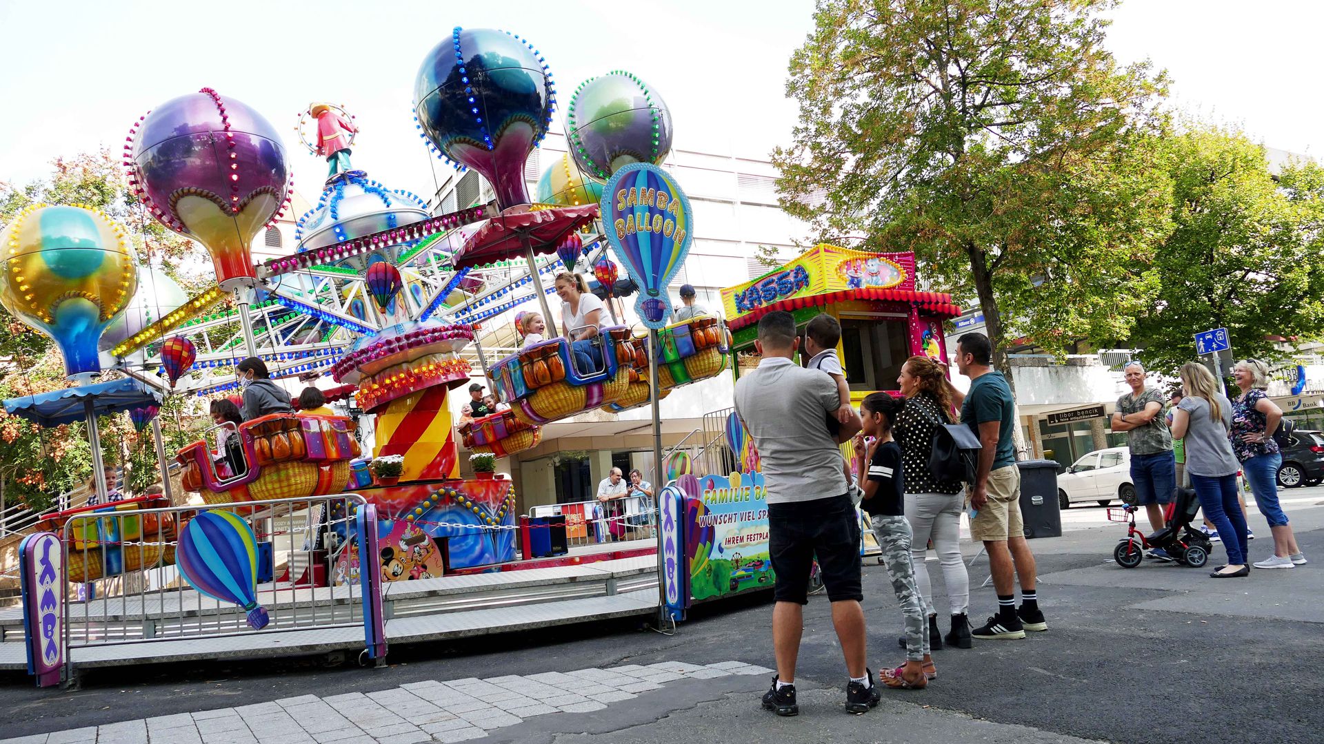 Mehrere Personen stehen vor einem Fahrgeschäft, das ein Karussell mit Heißluftballons ist.
