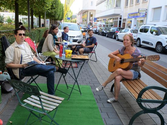 Mehere Personen sitzen gemütlich an Tischen in einer Parkzone an der Straße, eine Frau hat eine Gitarre in der Hand.
