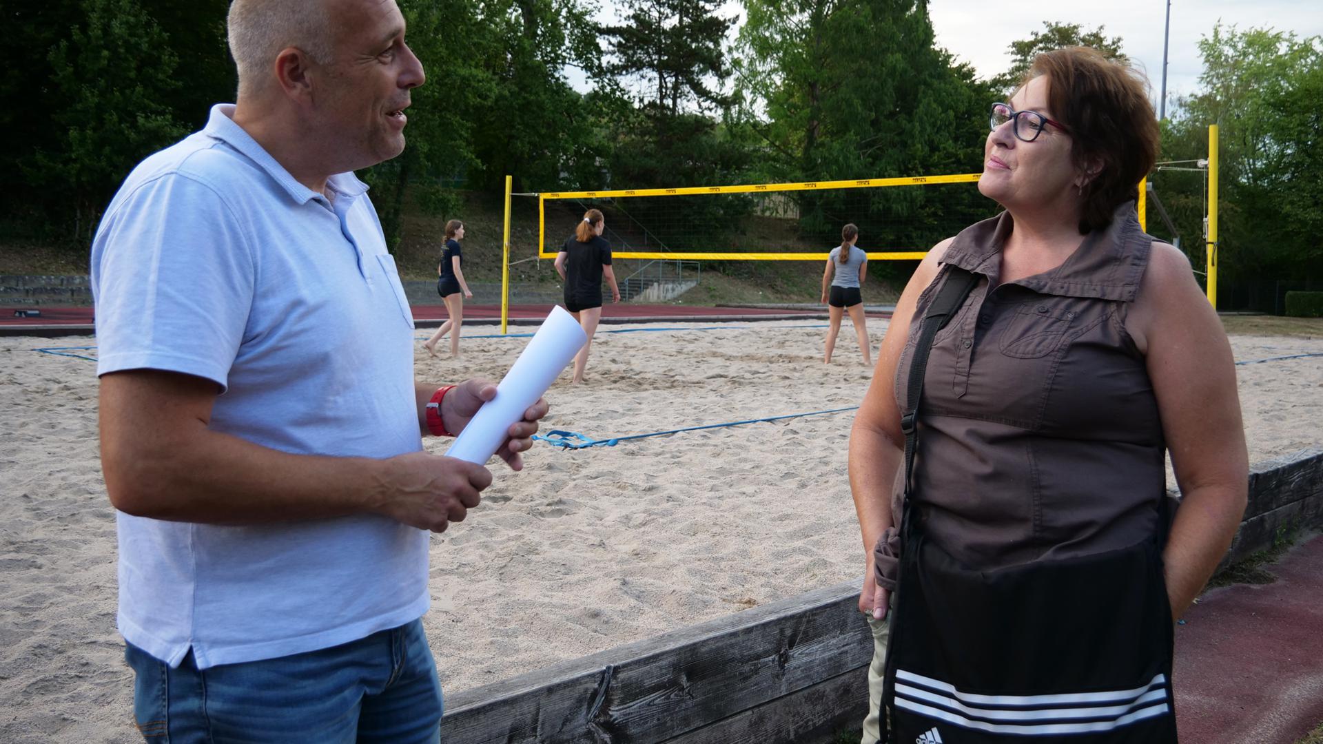 Ein Mann und eine Frau stehen vor einem Beachvolleyball-Feld und sprechen miteinander.