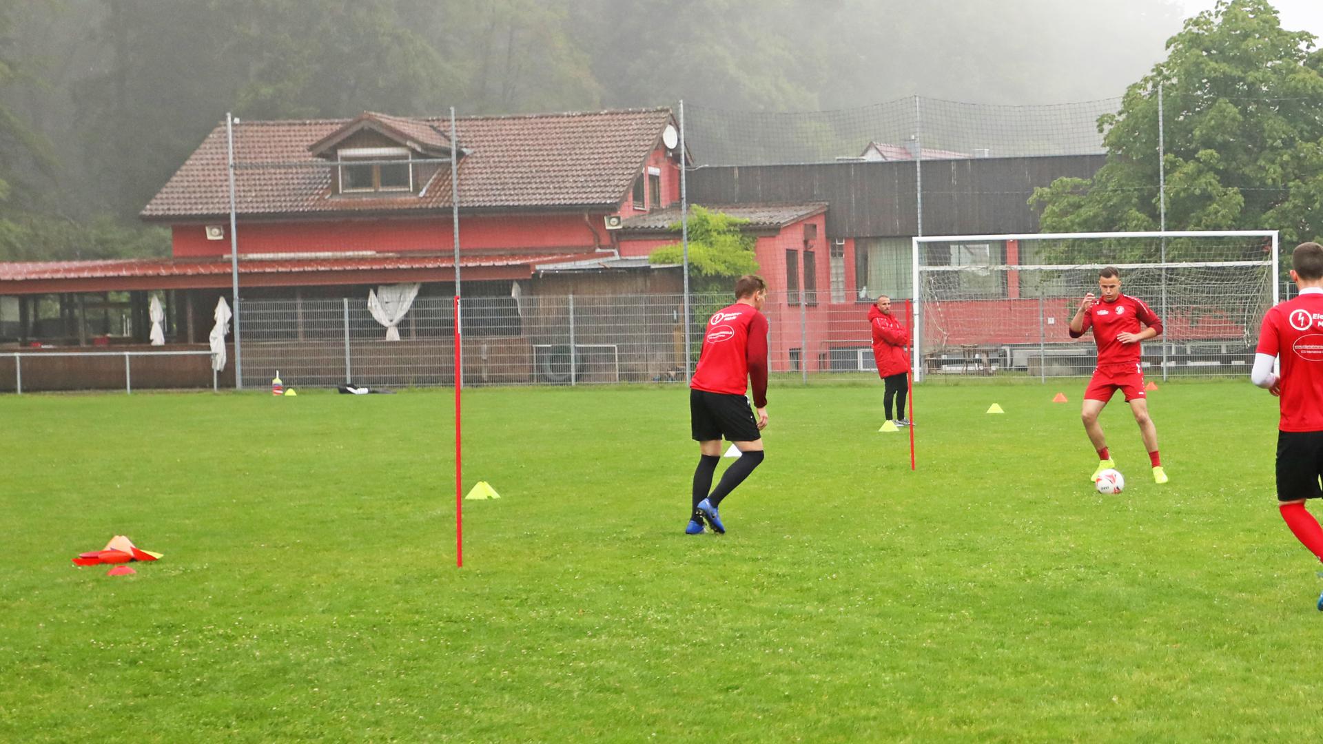Im Vordergrund spielen mehrere junge Männer Fußball auf einem grünen Rasenplatz, dahinter ragt das Vereinsheim auf.