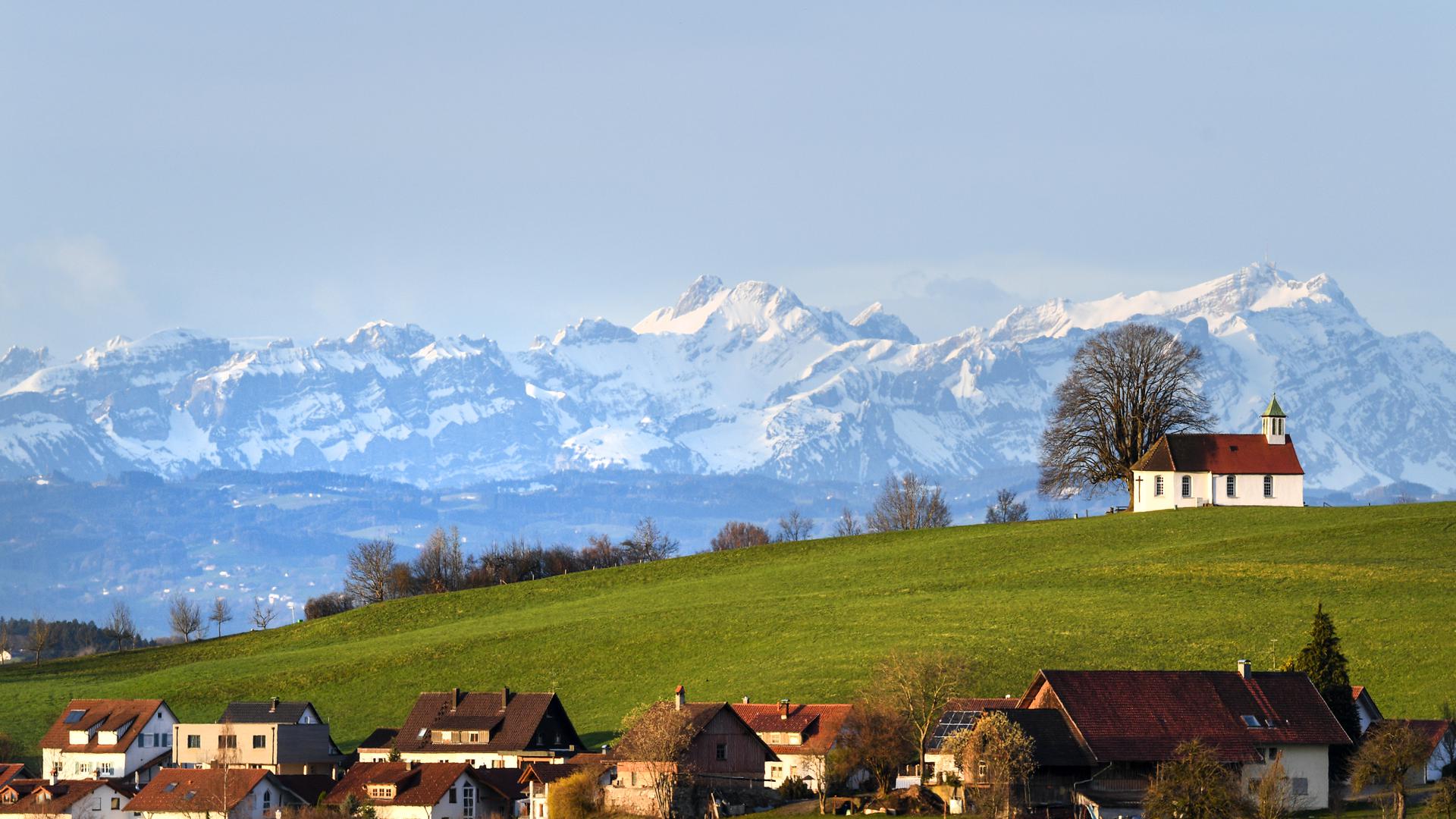 Die Morgensonne strahlt die Heilig-Kreuz-Kapelle auf dem Kapellenberg an, während im Hintergrund der Berg Säntis in der Schweiz zu sehen ist. 