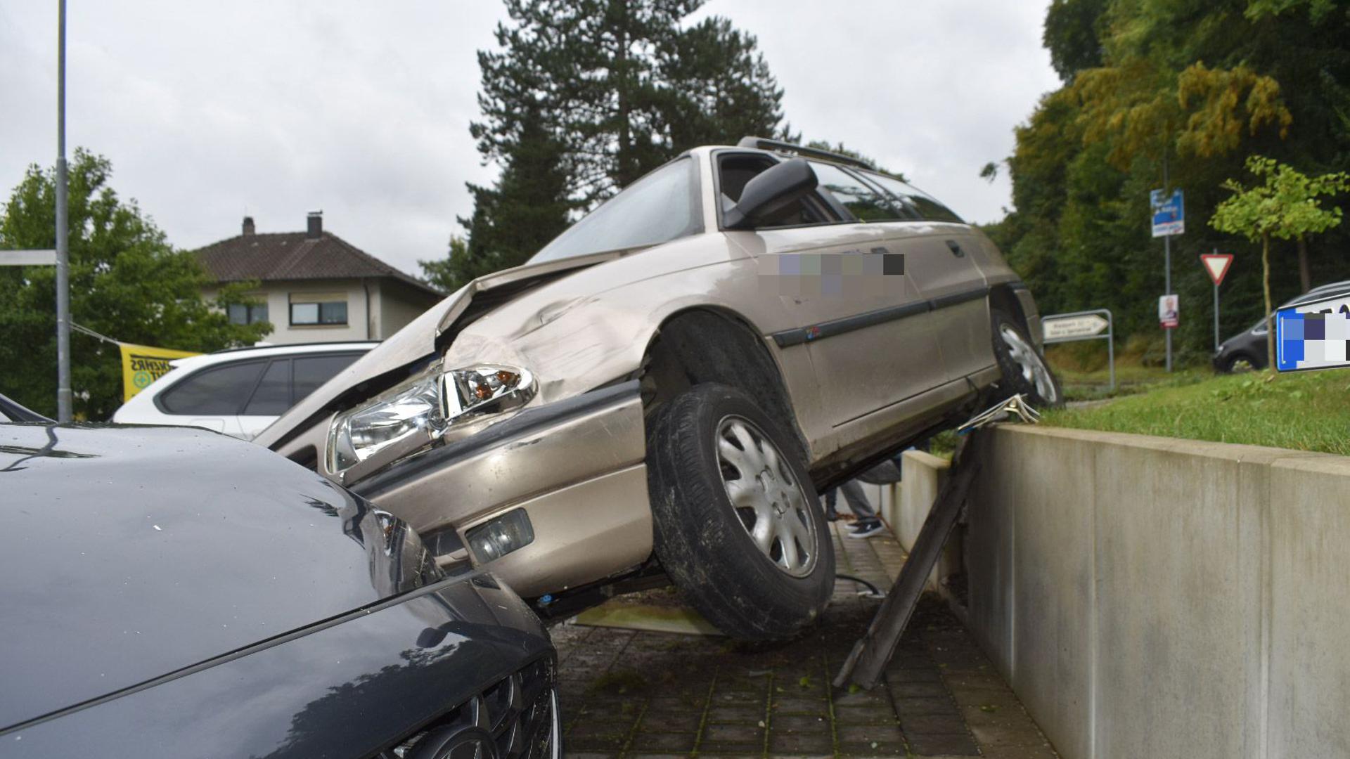 Ein graues Auto hängt zwischen einem Grünstreifen und einem geparkten Auto auf einem tieferliegenden Parkplatz in der Luft. 