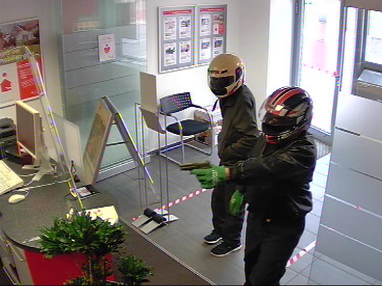 Zwei Männer stehen in einem Vorraum einer kleinen Bankfiliale, beide haben Helme auf, einer hält eine Pistole in der ausgestreckten Hand. 