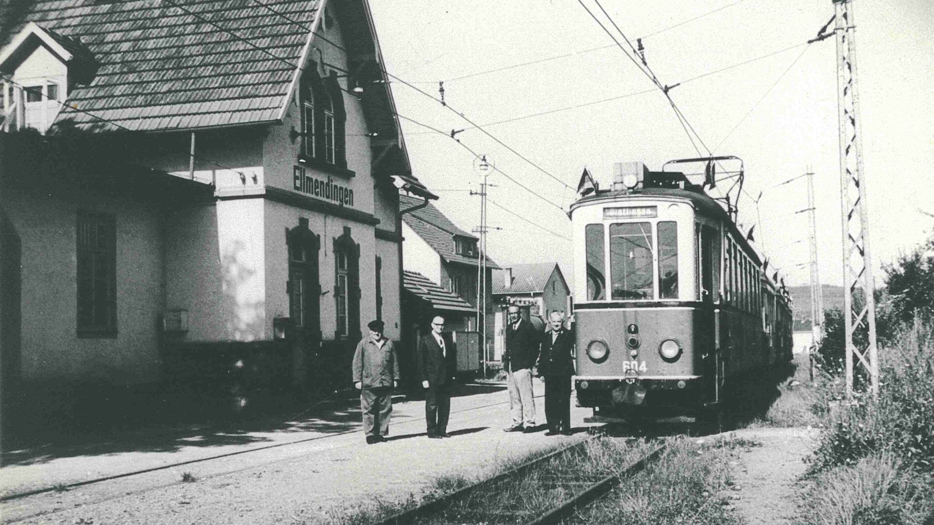 Eine Bahn steht auf den Gleisen vor dem Bahnhof Ellmendingen. Vier Männer stehen zwischen Bahn und Bahnhof. 