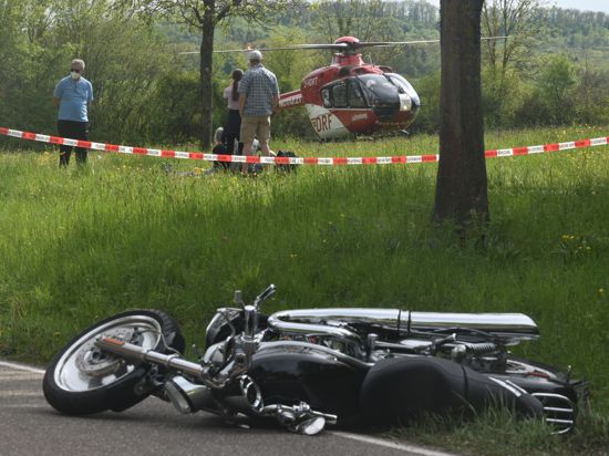 Ein schwarzes Motorrad liegt auf der Straße. Im Hintergrund steht ein Rettungshubschrauber auf einer Wiese.