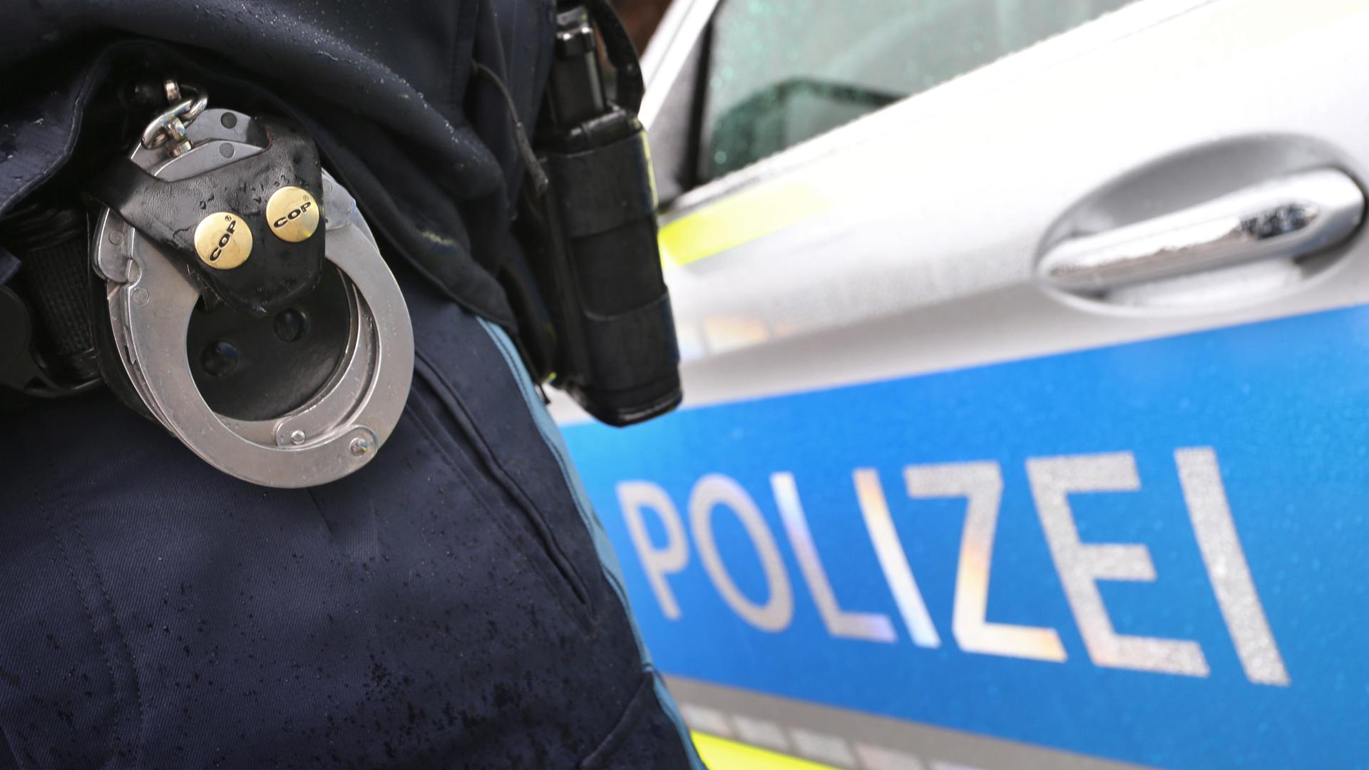 ILLUSTRATION - Ein Polizist mit Handschellen und einer Pistole am Gürtel steht vor einem Streifenwagen. Die bayerische Polizei fahndet derzeit nach 30 160 Verdächtigen oder Verurteilen. +++ dpa-Bildfunk +++