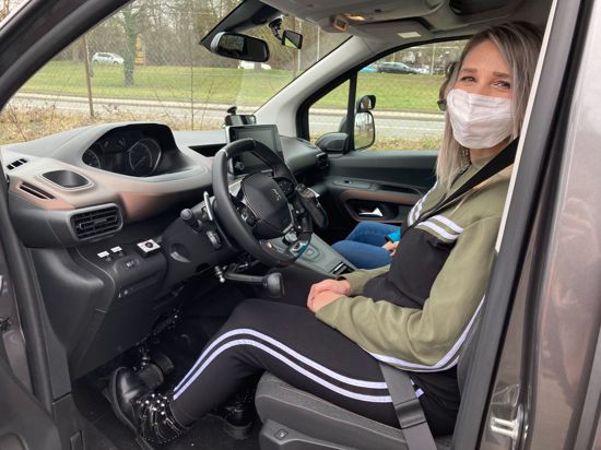 Eine Frau mit Mund-Nasen-Bedeckung sitzt am Steuer eines Autos und schaut durch die geöffnete Tür in die Kamera. 