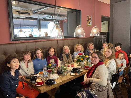 Geflüchtete Ukrainerinnen sitzen in einem Café an einem langen Tisch dicht beieinander und blicken in die Kamera. Sie lächeln müde. Das Peace Café in Pforzheim wird von der Organisation Golden Hearts organisiert.