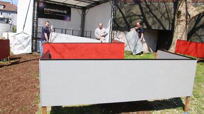 Drei Männer stehen vor einer Open-Air-Bühne, auf der das Wort „Kupferdächle“ zu lesen ist und bauen davor eine große grau-rote Box auf.
