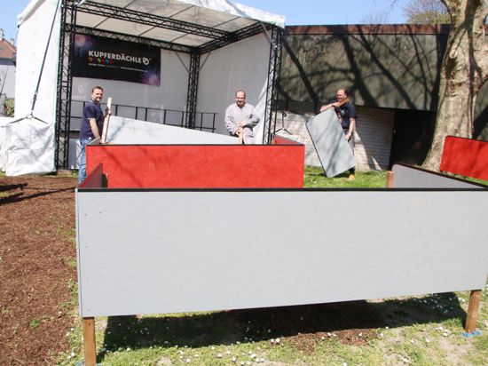 Drei Männer stehen vor einer Open-Air-Bühne, auf der das Wort „Kupferdächle“ zu lesen ist und bauen davor eine große grau-rote Box auf.