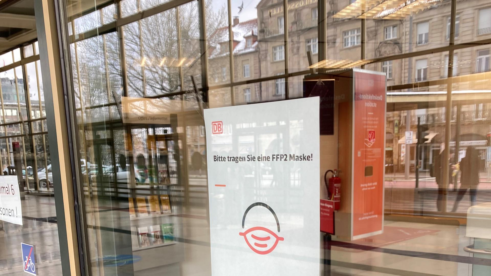 Maskenbitte statt Maskenpflicht: Auf einem Zettel im DB-Reisezentrum im Pforzheimer Hauptbahnhof steht „Bitte tragen Sie eine FFP2-Maske!“. In der Scheibe spiegelt sich der Schlosskeller.