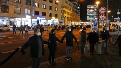 Teilnehmer der Corona-Demonstration ziehen am Pforzheimer Stadttheater vorbei, im Vordergrund halten sich Bürger bei einer Menschenkette für die Demokratie an den Händen.
