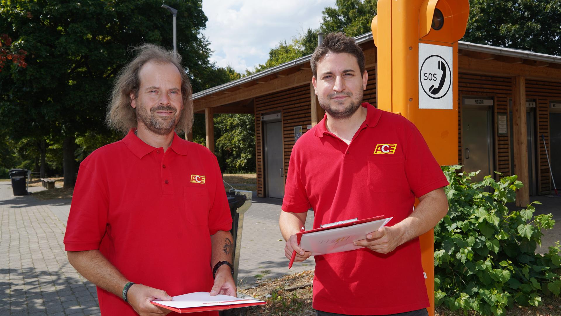 Michael Walter (links) und Elias Schempf vom Auto Club Europa stehen vor einem Toilettenhäuschen mit Notrufsäule