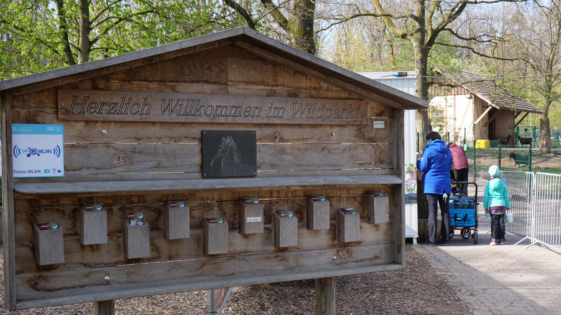 Ein Holzhäuschen mit der Aufschrift „ Herzlich Willkommen im Wildpark“ steht im Vordergrund, im Hintergrund sind Menschen und ein Gehege mit Tieren.