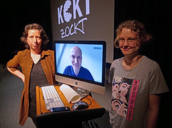 Zwei Frauen neben einem Rechner, auf dem ein Mann zu sehen ist. Im Hintergrund projiziert der Titel „Koki zockt“. 