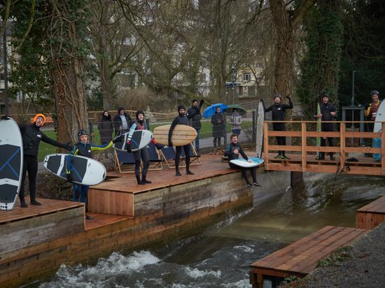  Menschen im Neopren-Anzug stehen an einem Kanal, schauen in die Kamera und haben Surf-Boards dem Arm. 