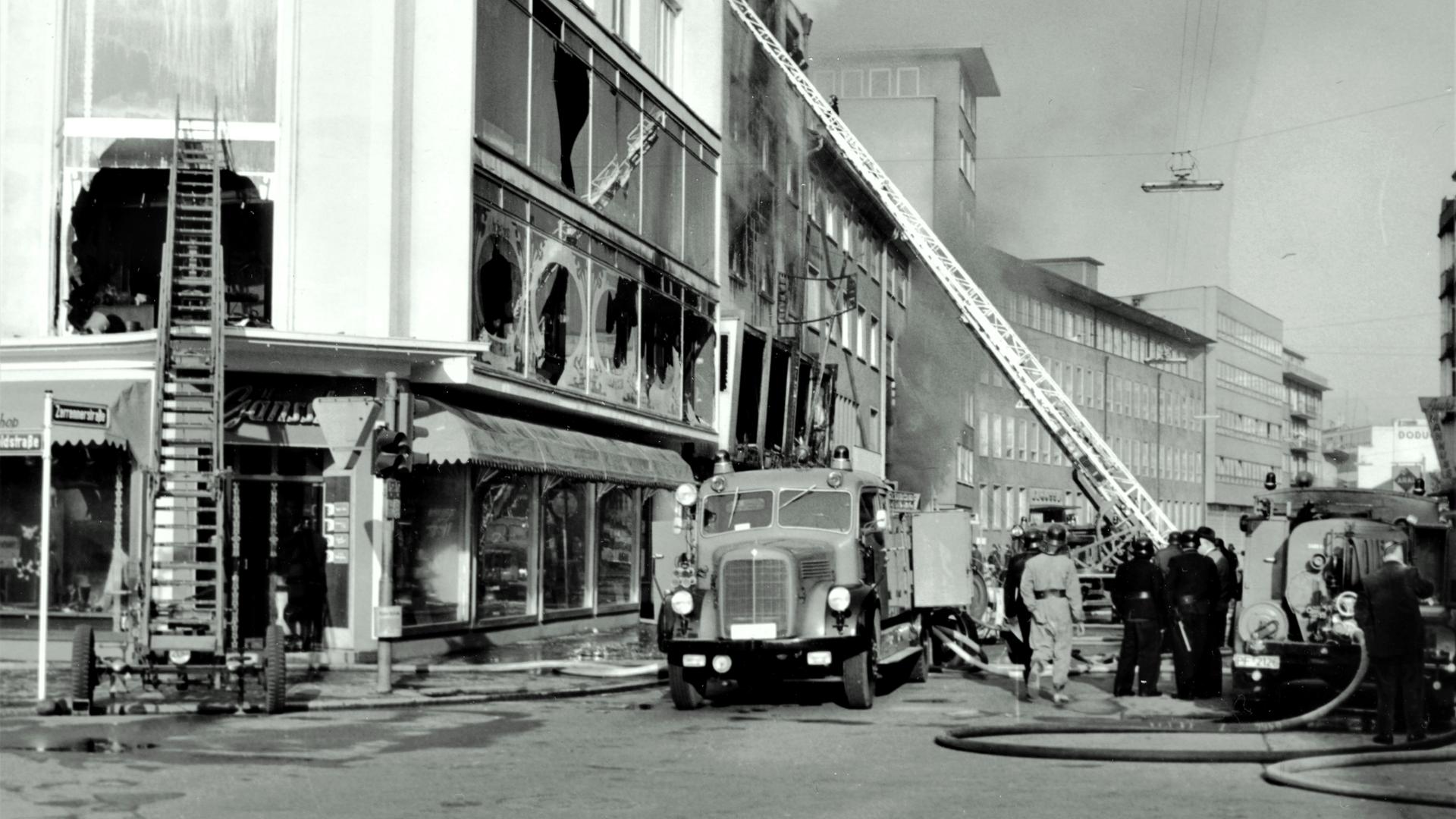 Schwarz-weiß-Foto eines Brands: Feuerwehr mit Leitern vor einem Haus mit geborstenen Fensterscheiben.